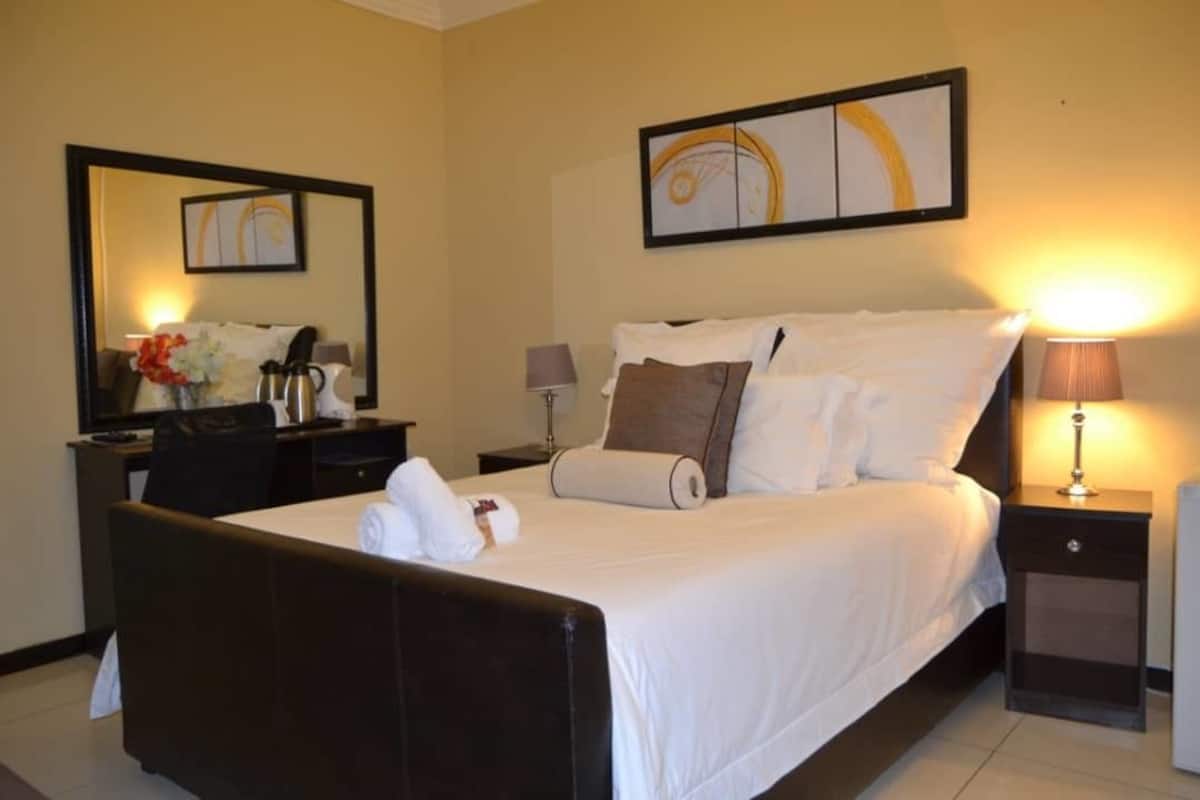 A hotel room with a large bed, a desk with a chair, a nightstand with a lamp, and a wall-mounted artwork.