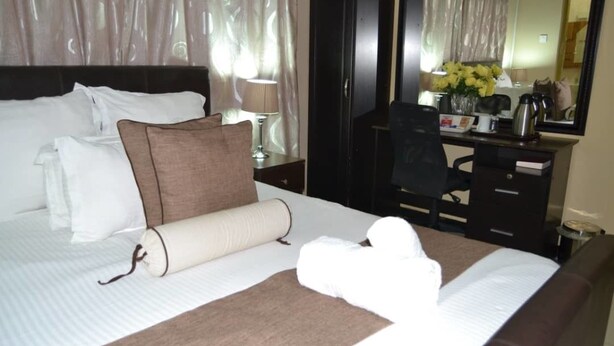 A neatly made bed with white and brown bedding, a desk with a lamp, and a mirror reflecting a room with curtains. - Jasper Guest House (Selebi-Phikwe)