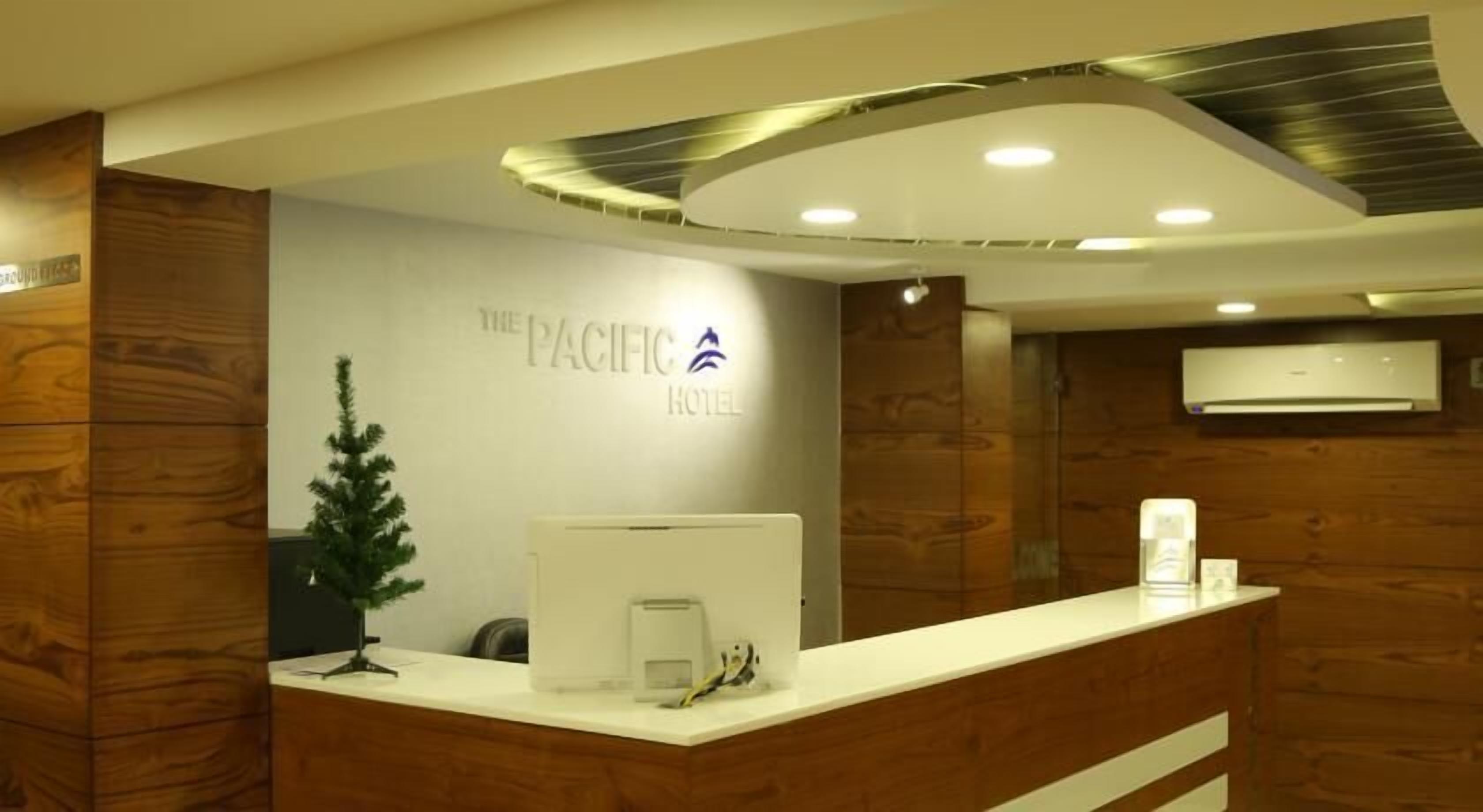 A reception desk with a wooden panel design, a small potted plant, and a digital screen. The reception area has a logo for 'The Pacific Hotel' on the wall.