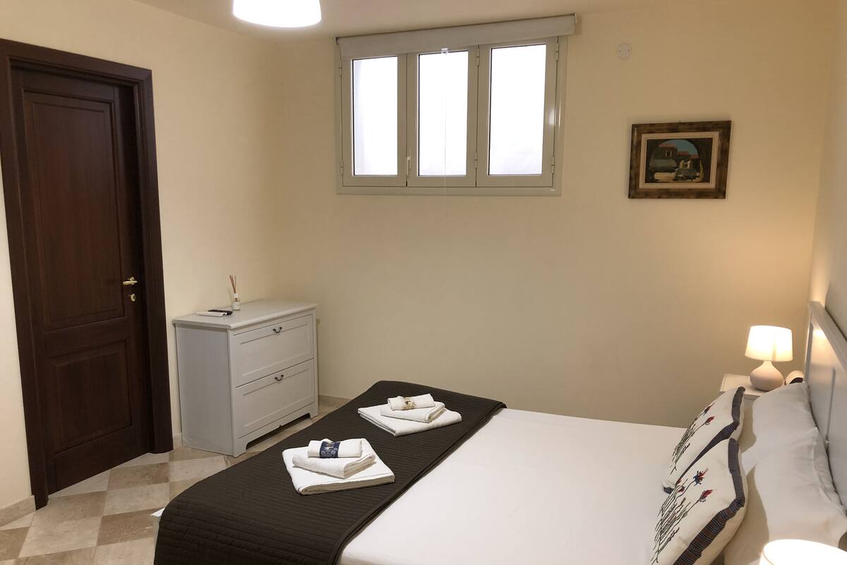 Quarto bem arrumado com cama, abajures, janela, porta e cômoda pequena.