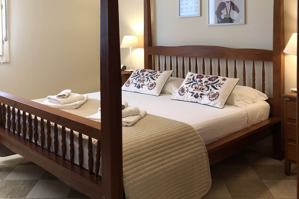 Uma cama de dossel com estrutura de madeira, roupa de cama branca e almofadas decorativas. Há duas obras de arte penduradas na parede e uma mesa de cabeceira com um abajur.