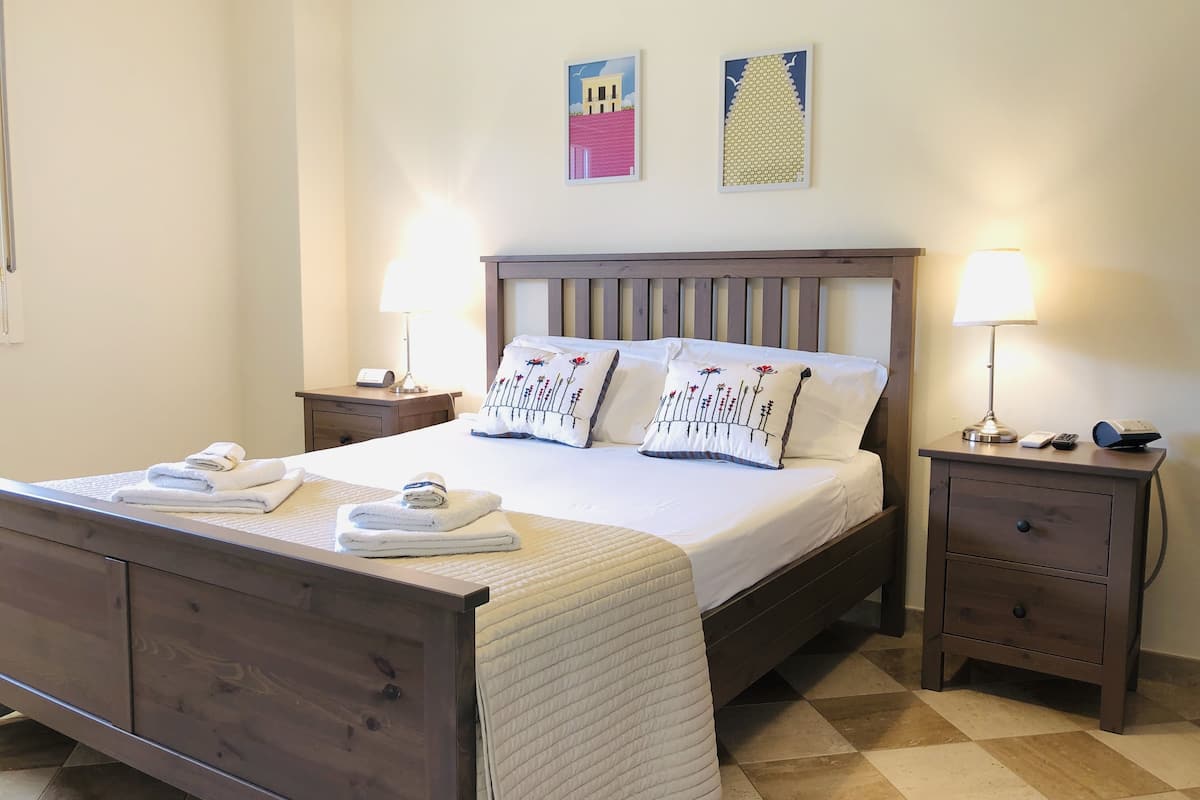 Uma cama bem arrumada com roupa de cama branca e almofadas decorativas, uma cabeceira de madeira, duas mesas de cabeceira com abajures e um piso xadrez.