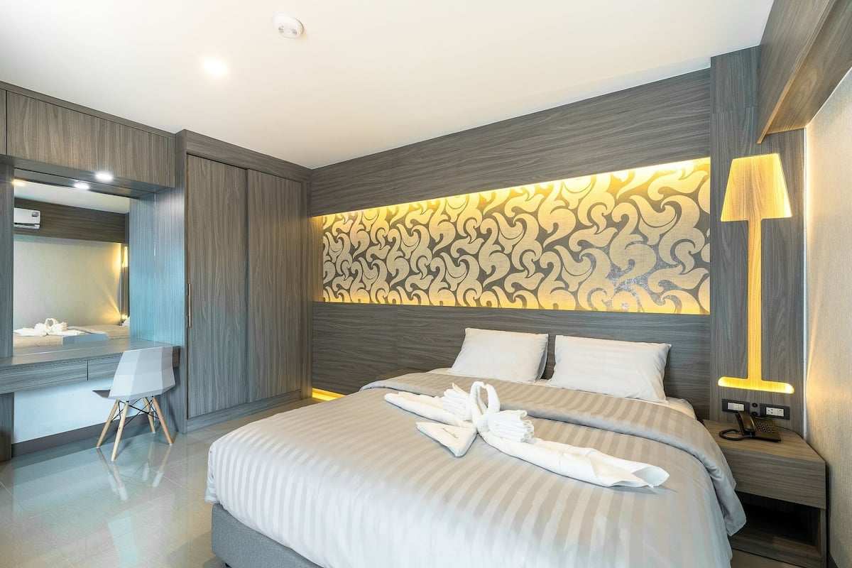 A modern hotel room with a large bed, a desk with a chair, a vanity with a mirror, and a patterned wall behind the bed.