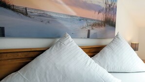 En säng med vita kuddar och en tavla som föreställer en strandscen, placerad på väggen bakom sängen.