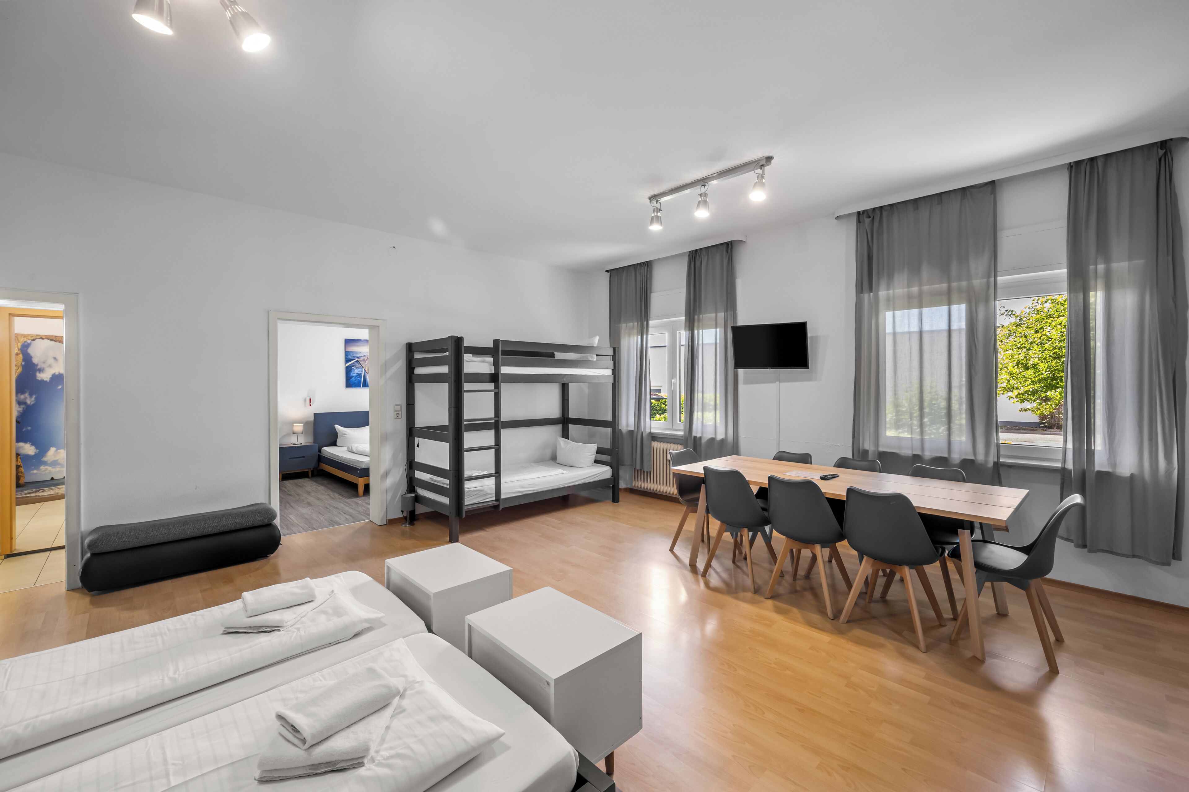 Ein modernes Hotelzimmer mit Etagenbetten, einem Esstisch mit Stühlen, einem Fernseher und großen Fenstern mit Vorhängen.