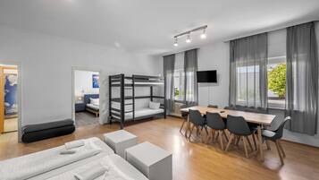 Una habitación de hotel moderna con literas, una mesa de comedor con sillas, un televisor y ventanas grandes con cortinas.