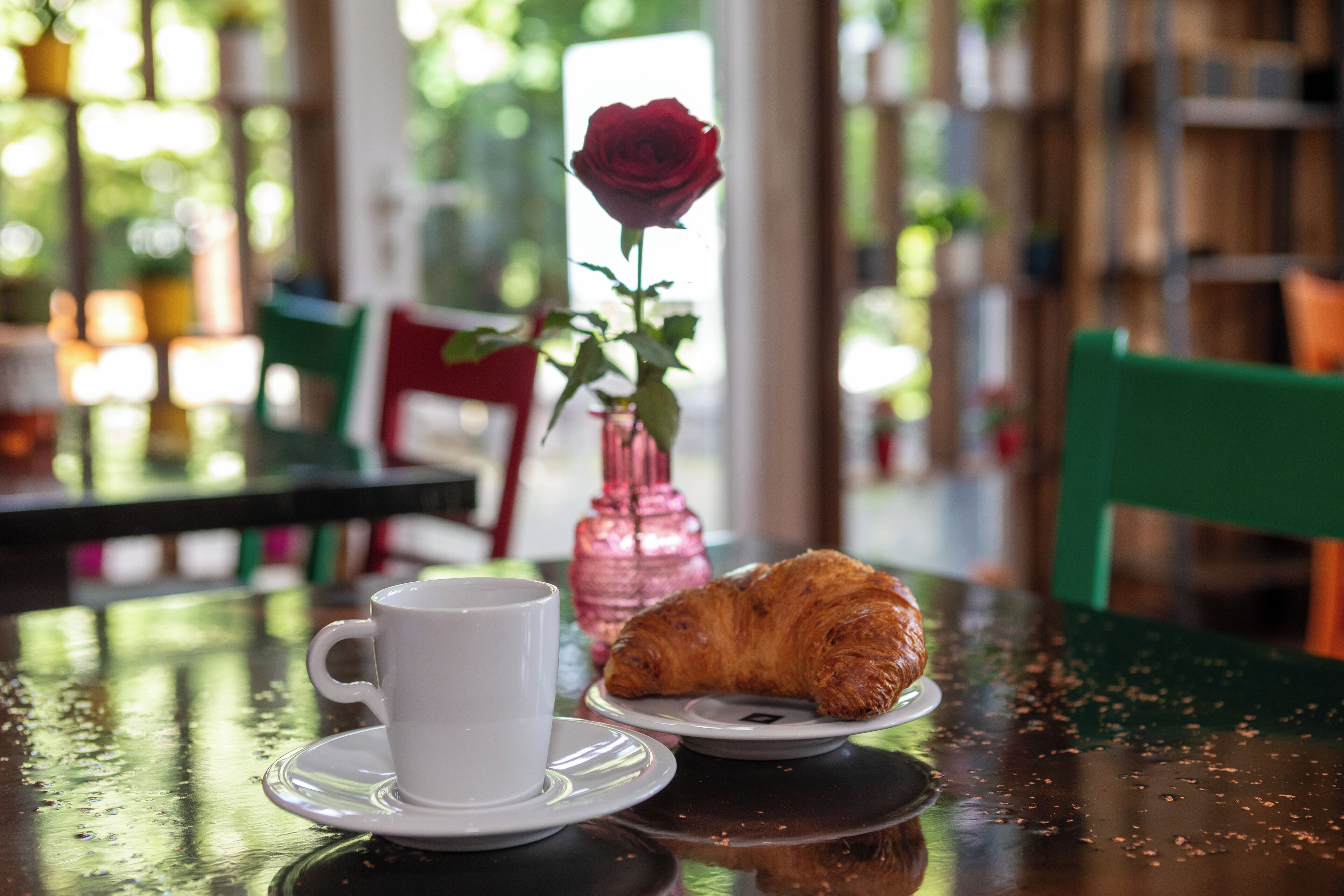 A table with a white cup and saucer, a plate with a croissant, and a pink vase with a rose.