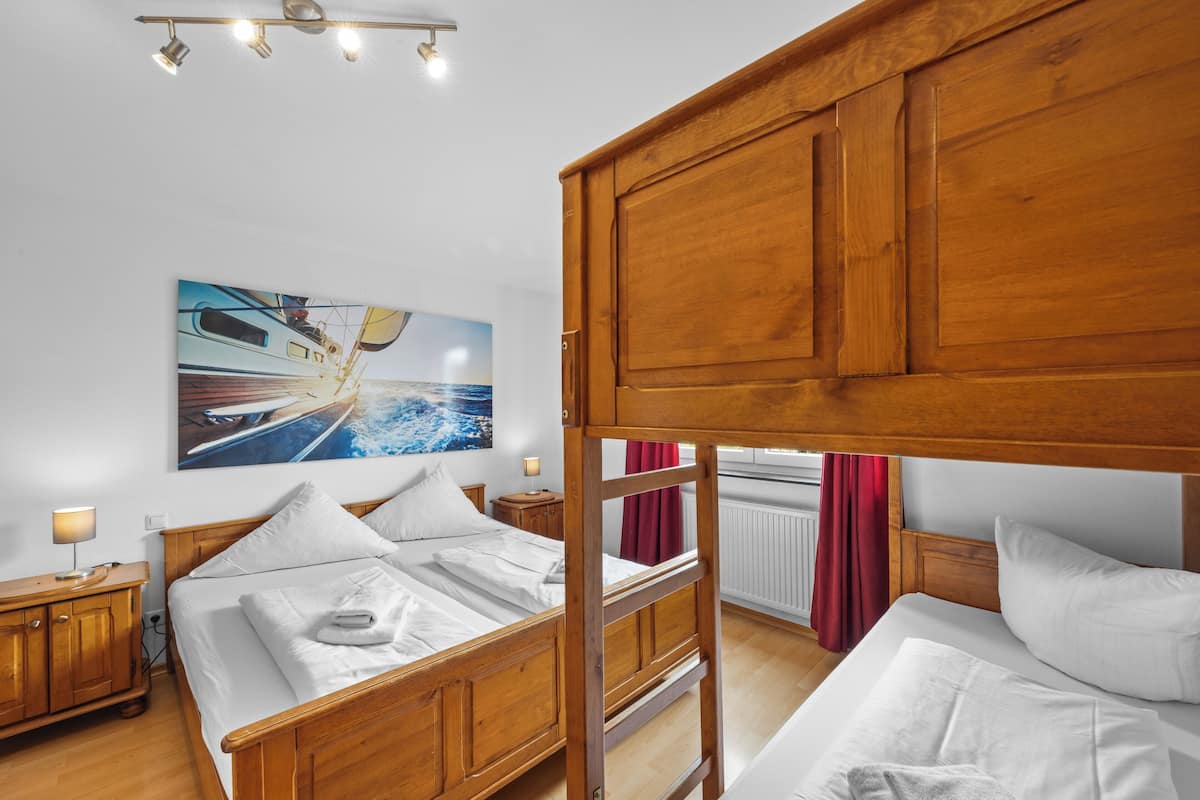 Une chambre avec un lit superposé, une tête de lit en bois, un tableau représentant un bateau accroché au mur, une table de chevet avec une lampe et une fenêtre avec des rideaux.