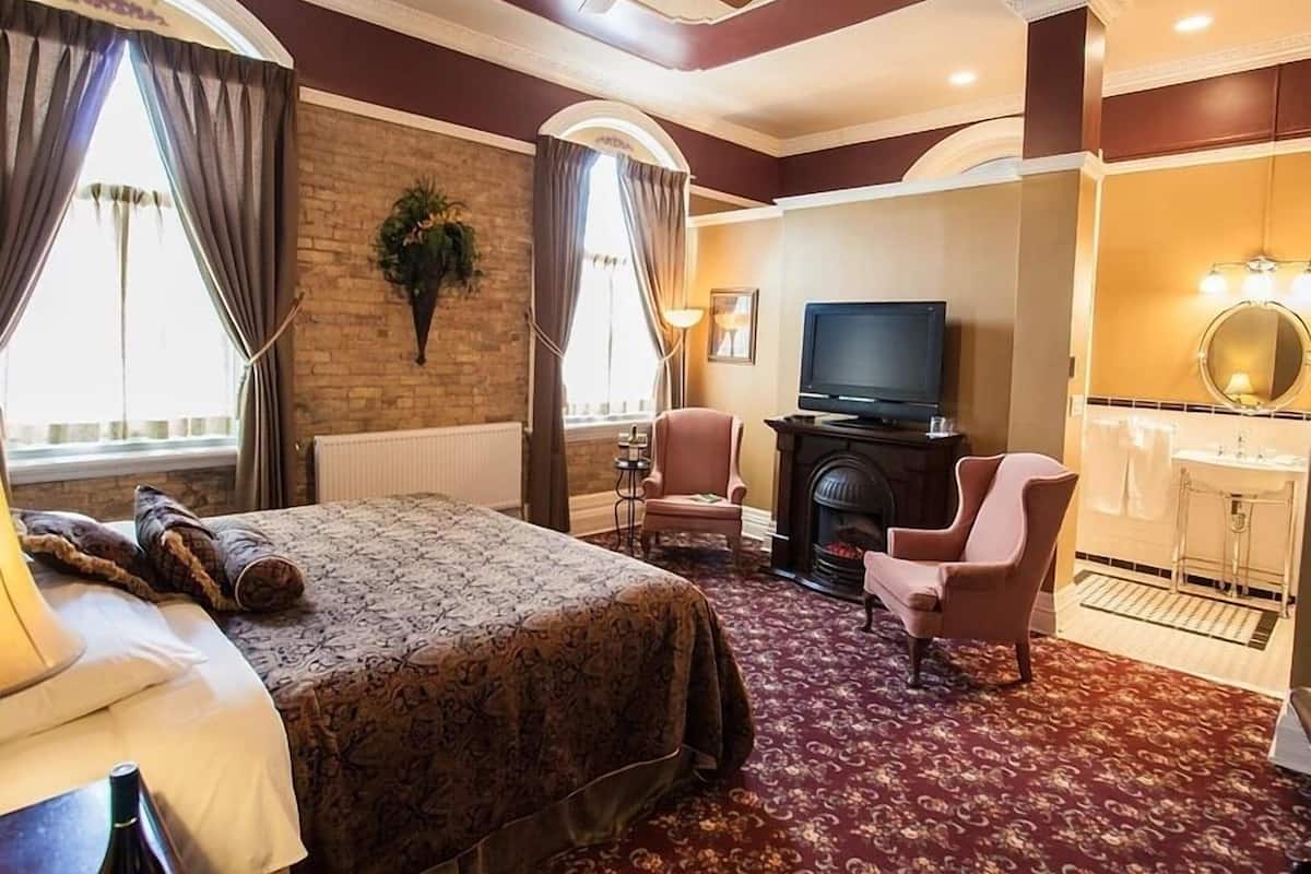 A hotel room with a bed, a television, a fireplace, a desk, and a bathroom with a mirror and sink.