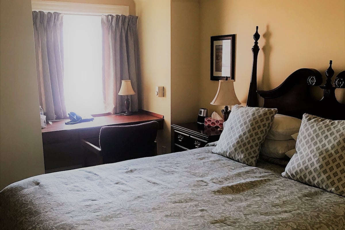 A bedroom with a bed, a desk, a chair, a lamp, a nightstand, and a window with curtains.
