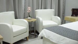 Ein Hotelzimmer mit einem Bett, zwei weißen Ohrensesseln, einem kleinen runden Tisch mit Blumenvase und einem Nachttisch mit Lampe.