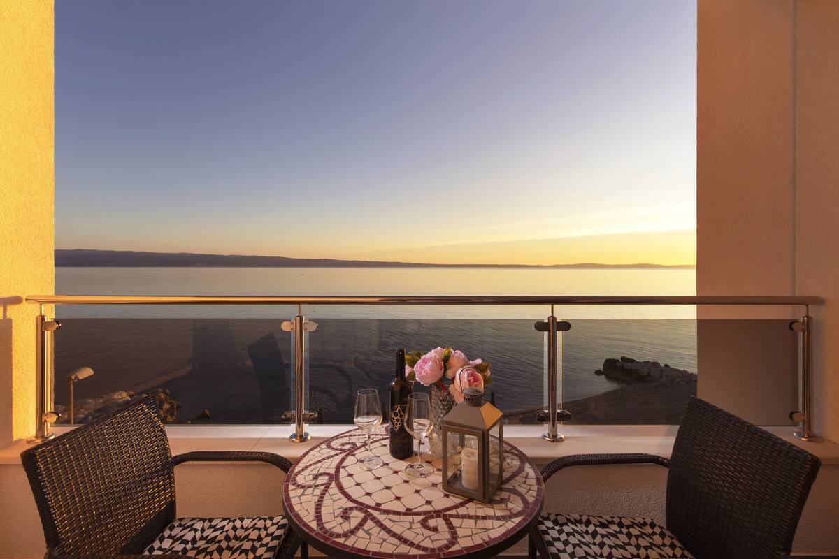 A balcony with a table set for two, featuring a view of the sea and a sunset.