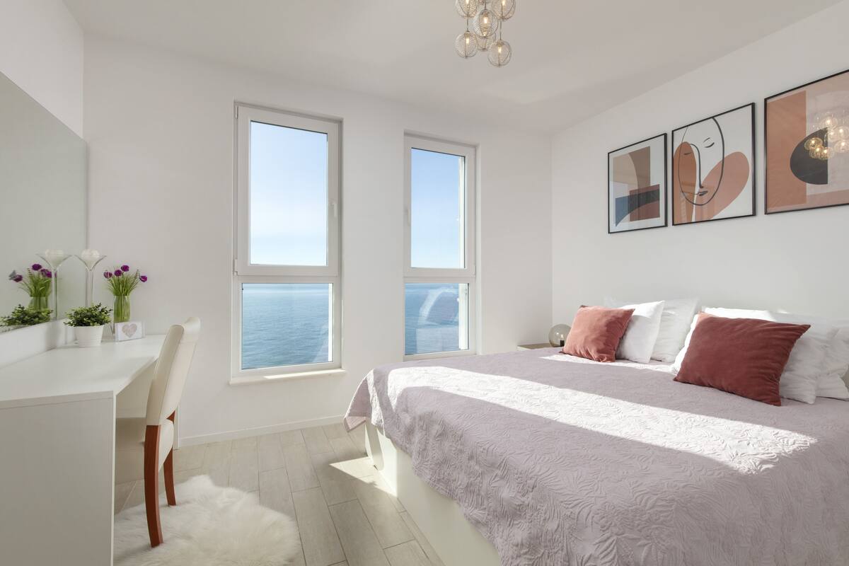 A modern bedroom with a large bed, a desk, and a view of the sea through two windows.