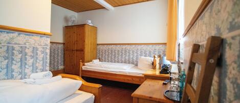 Una habitación pequeña y acogedora con dos camas individuales, un armario de madera, una mesita con una lámpara y una silla.