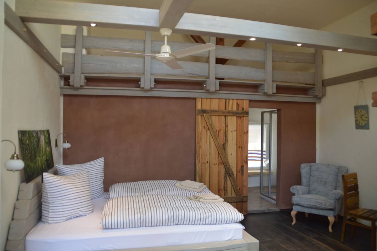 Une chambre à coucher avec un lit, une chaise, un ventilateur de plafond et une porte en bois.
