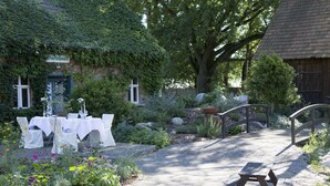 A garden setting with a picnic table and tables set up for dining, surrounded by lush greenery and a rustic building with a sign reading "Backhaus". - Hotel Kolonieschänke (Burg (Spreewald))