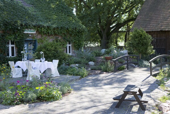 A garden setting with a picnic table and tables set up for dining, surrounded by lush greenery and a rustic building with a sign reading "Backhaus". - Hotel Kolonieschänke (Burg (Spreewald))