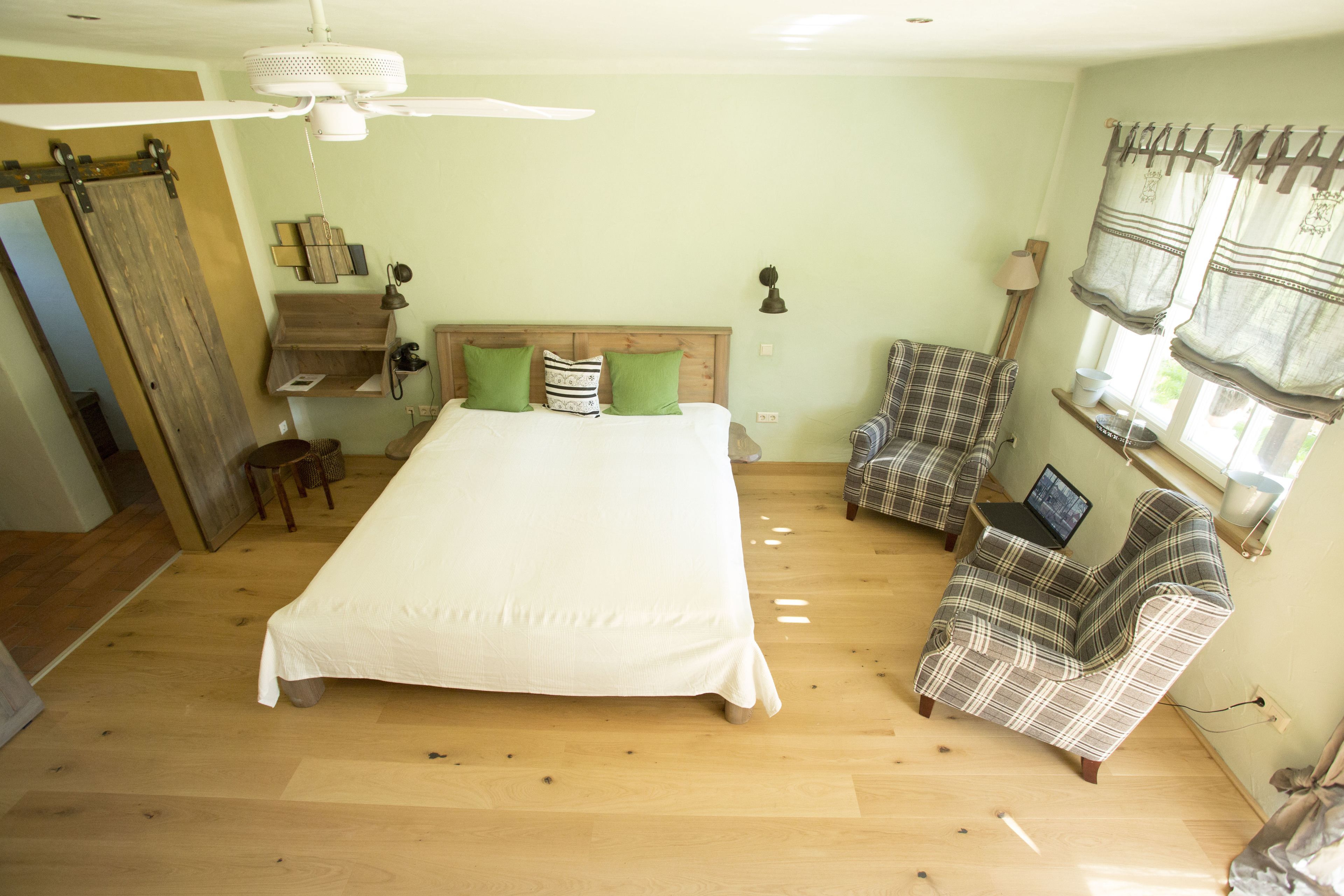 Un dormitorio con una cama grande, piso de madera, ventilador de techo, un sillón a cuadros y un ordenador portátil sobre una mesita.