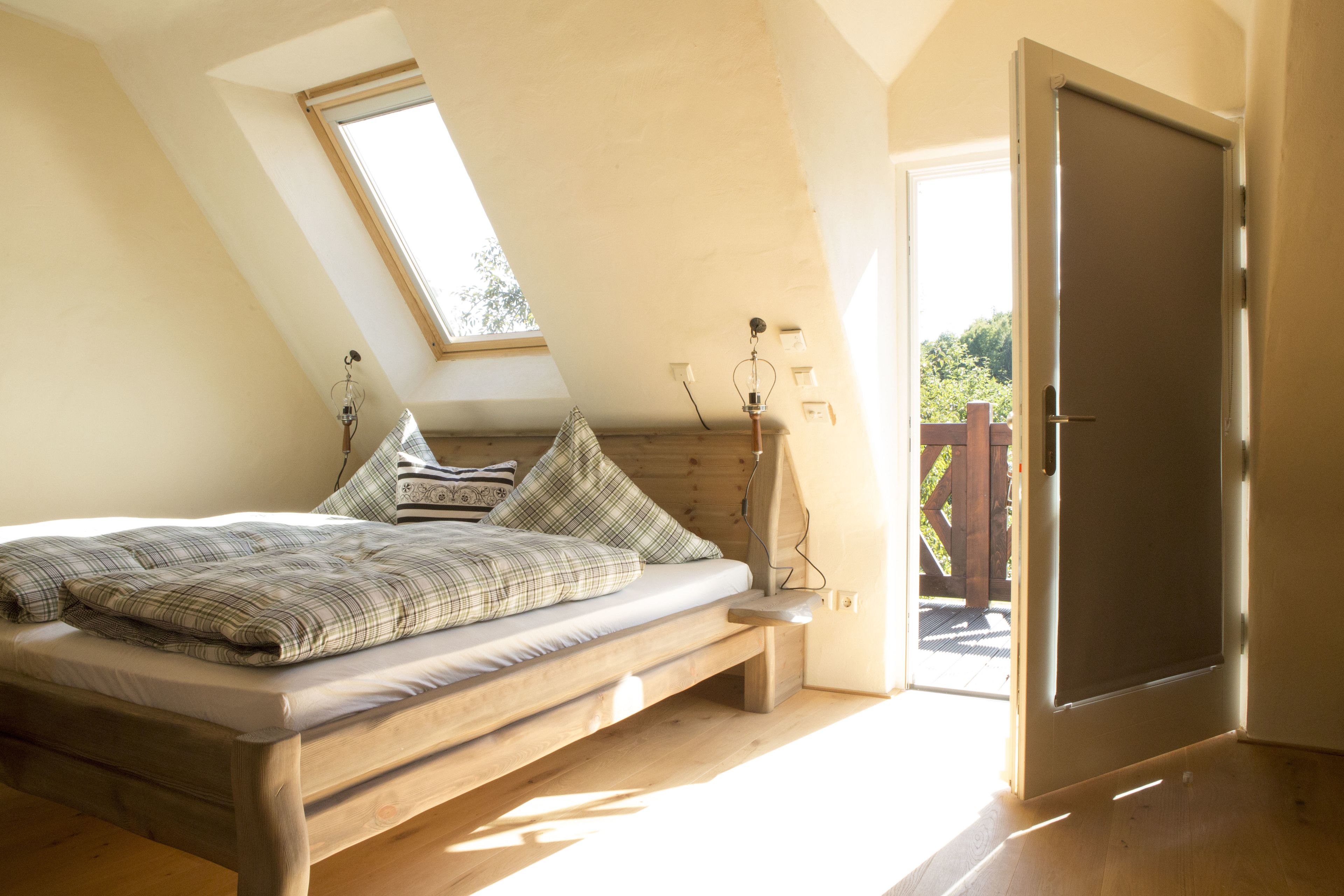 Ein Schlafzimmer mit schrägverdeckter Decke, einem hölzernen Bett, einem Fenster und einer zur Außenseite führenden Tür.