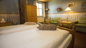Una habitación con dos camas, un armario de madera, un sillón verde, una mesita con lámpara y una ventana con cortinas.