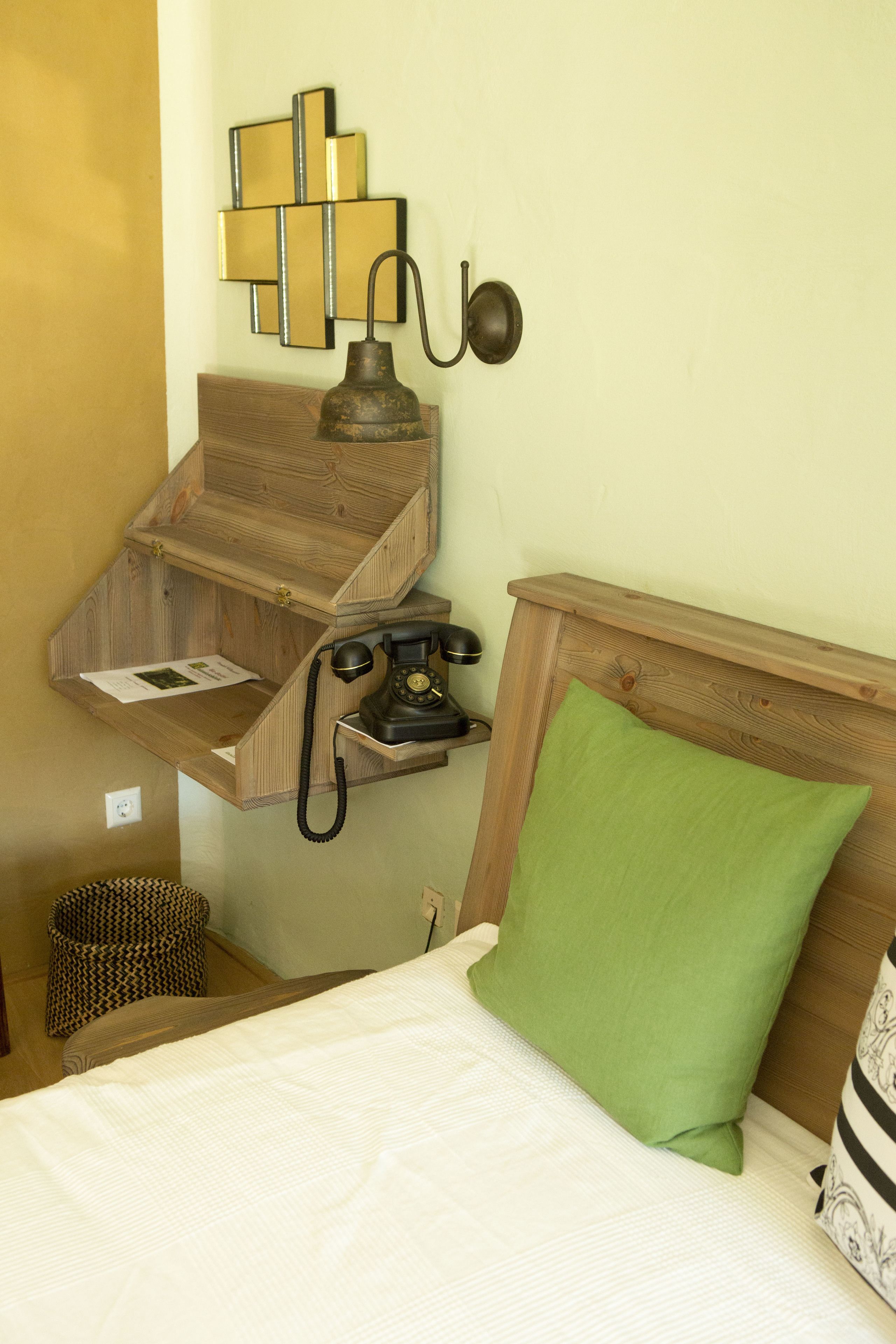 Quarto com cabeceira de madeira, travesseiro verde, telefone antigo e luminária fixada na parede.