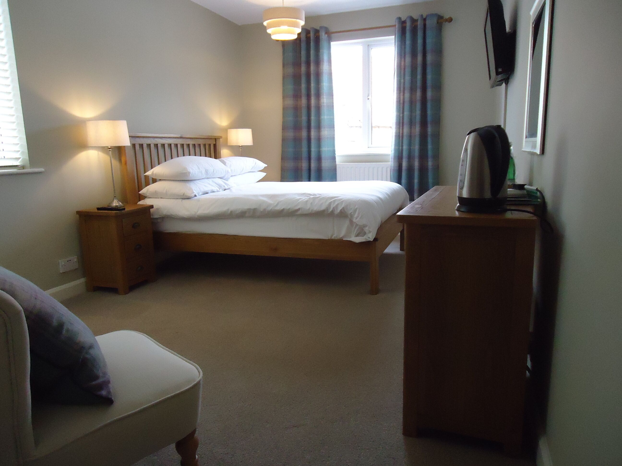 A bedroom with a bed, bedside tables, a chair, a desk, a radiator, a window with curtains, and a wall-mounted television.