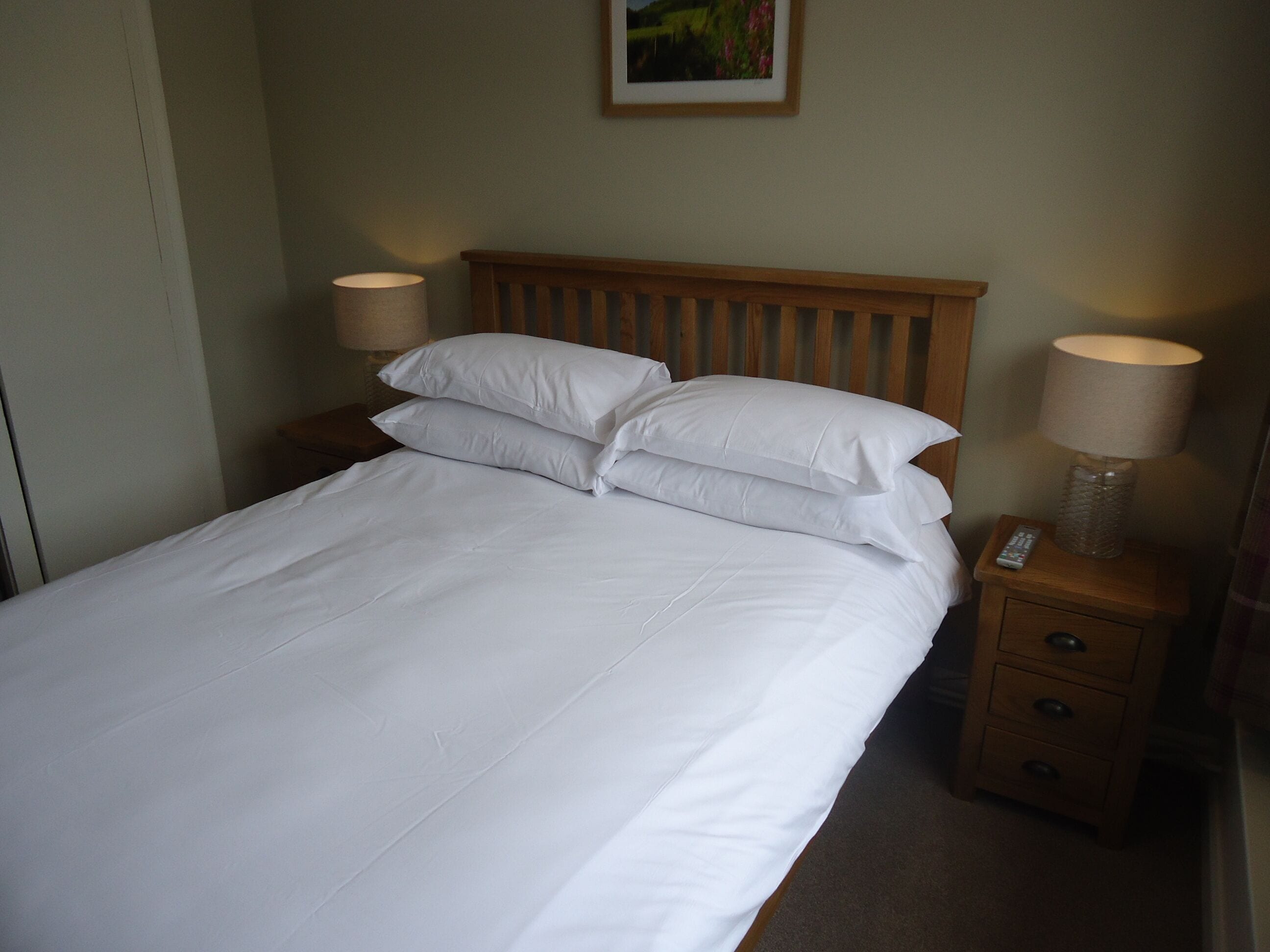 A neatly made bed with white linens and pillows, a wooden headboard, two bedside tables with lamps, and a small drawer.