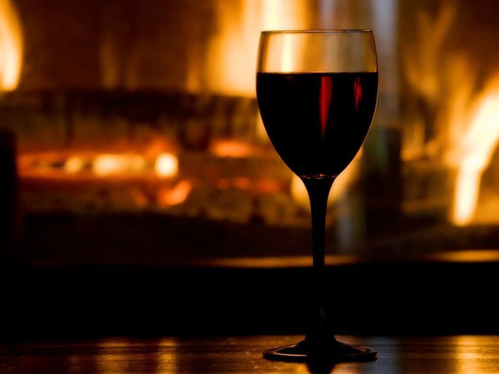 Een glas rode wijn op een houten oppervlak, met een onscherpe achtergrond van een warm verlichte kamer.
