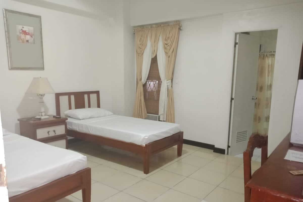 A hotel room with two single beds, a bedside table with a lamp, a small cabinet, and a window with curtains.