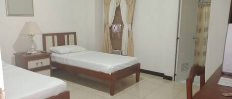 A hotel room with two single beds, a bedside table with a lamp, a small cabinet, and a window with curtains.
