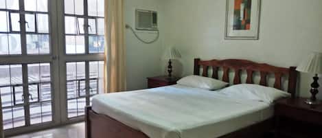 Una habitación con cama, ventana con cortinas, aire acondicionado de pared y un cuadro enmarco.