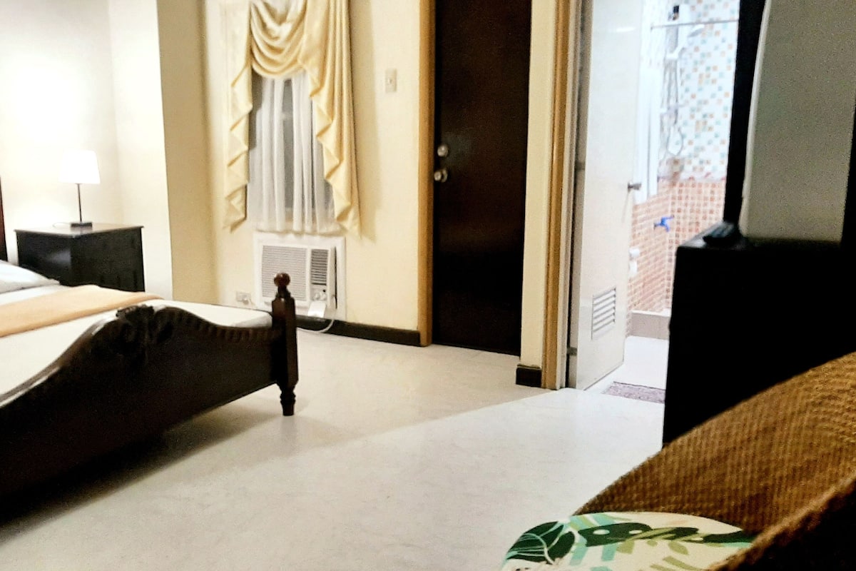 A hotel room with a bed, a nightstand, a lamp, a window with curtains, and a door leading to a bathroom.