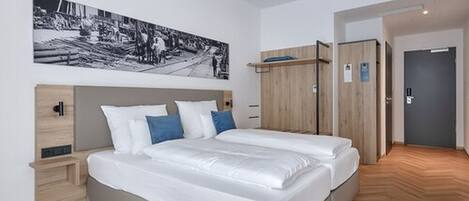A modern hotel room with a large bed, a wooden wardrobe, a small bedside table, and a wall-mounted artwork.