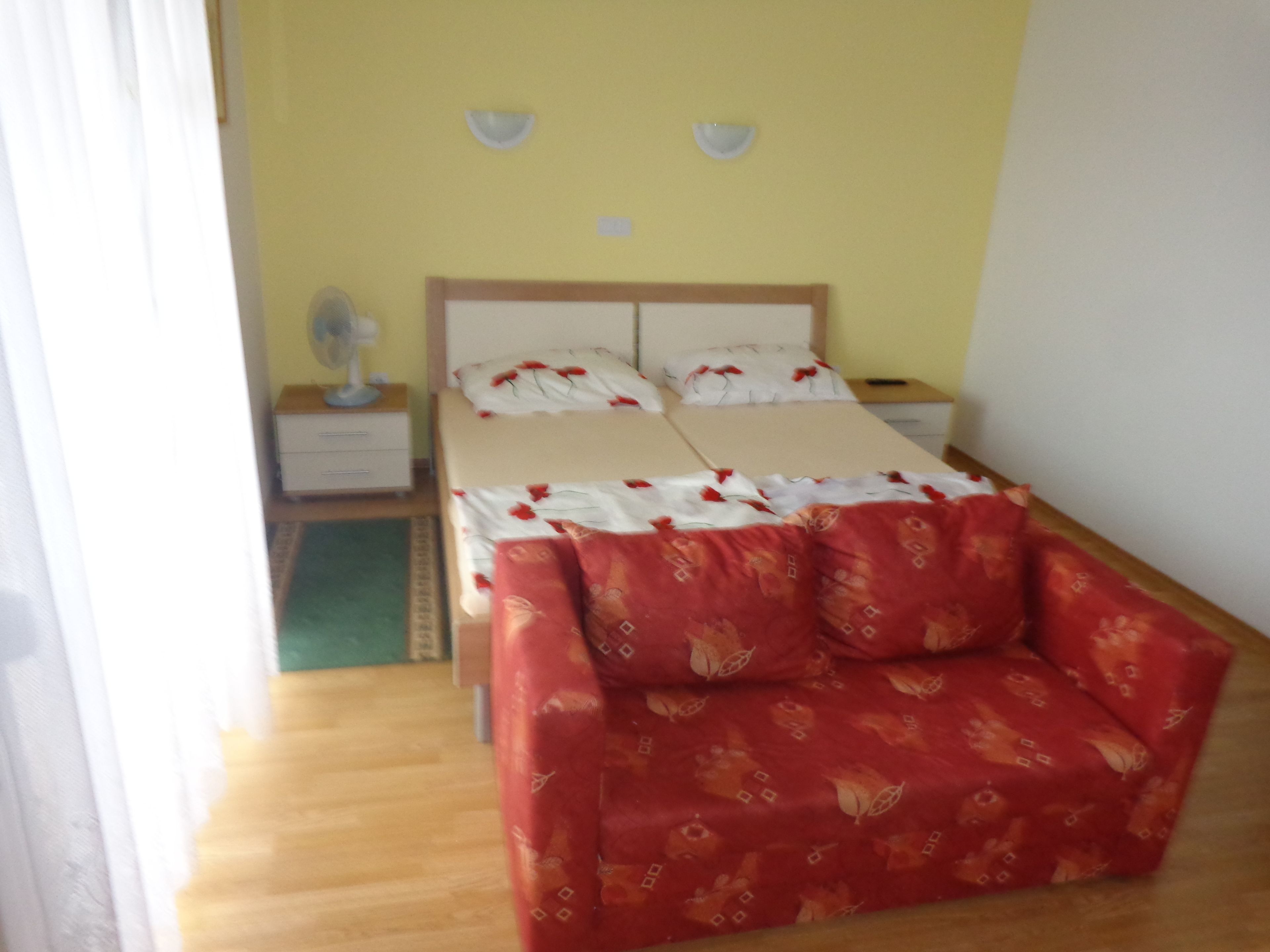Una cama doble con ropa de cama floral, una mesita de noche con lámpara, y un sofá rojo en una habitación con paredes amarillas.