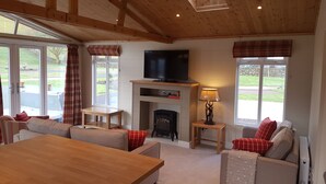 A spacious living room with a wooden ceiling, a fireplace, a television, and large windows. - Hendre Rhys Gethin (Betws-Y-Coed)