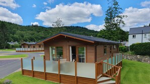 A wooden cabin with a glass balcony, a stone staircase, and a view of a road and forested hills. - Hendre Rhys Gethin (Betws-Y-Coed)