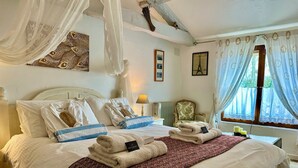 Een netjes opgemaakt bed met witte lakens, een blauw-wit gestreept kussen en een rode gebogen plaid. Er staat een klein tafeltje met een lamp en een decoratief object.