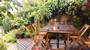 Een houten terras met een tafel en stoelen, omgeven door weelderige begroeiing en bloemen.