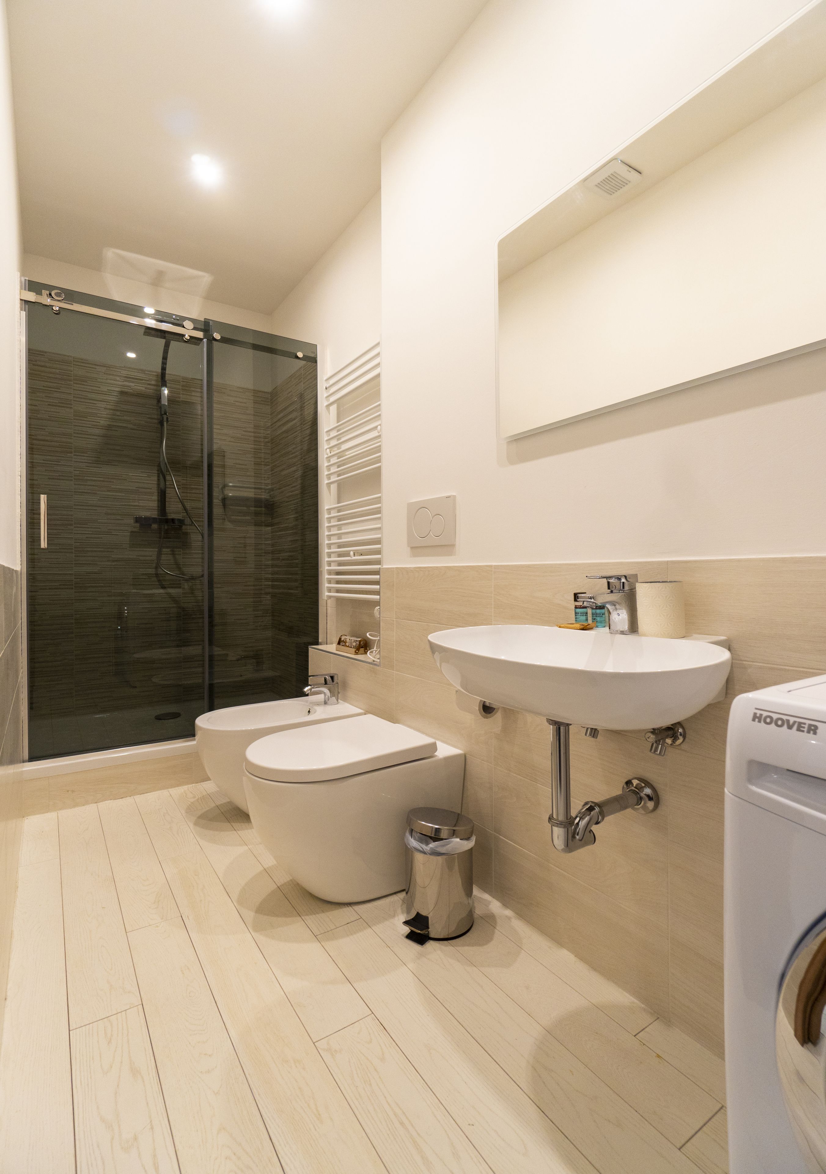 Uma casa de banho moderna com chuveiro envolto em vidro, sanita, bidé, lavatório e máquina de lavar roupa integrada.