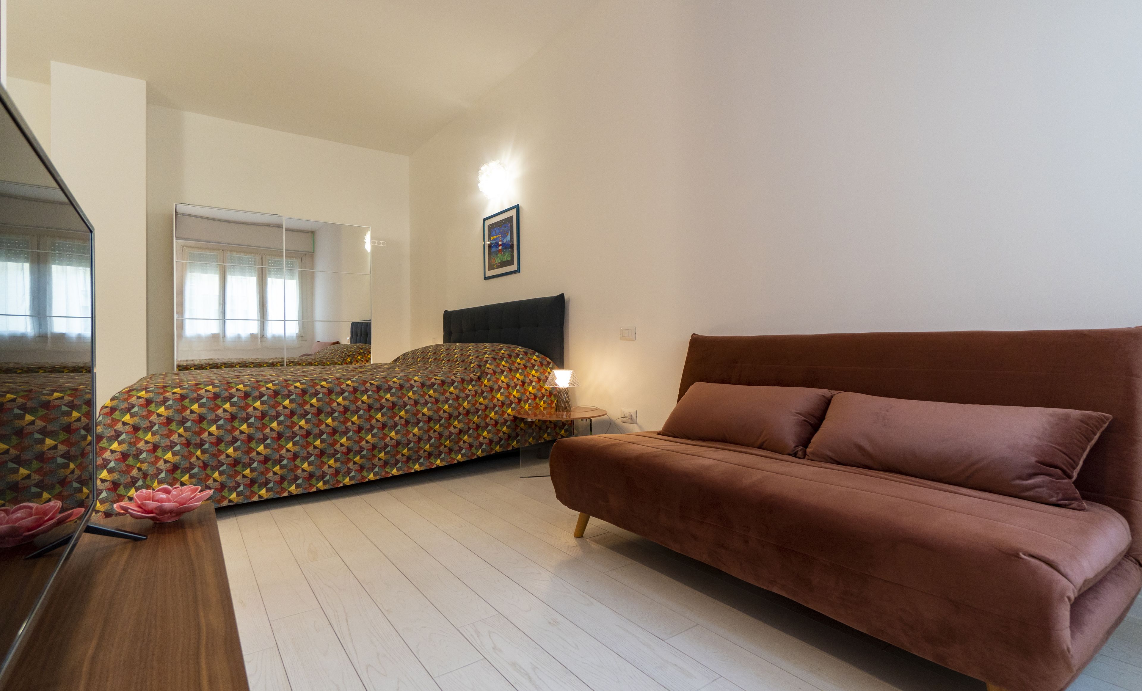 A bedroom with a bed, a brown sofa, a bedside table, and a mirror.