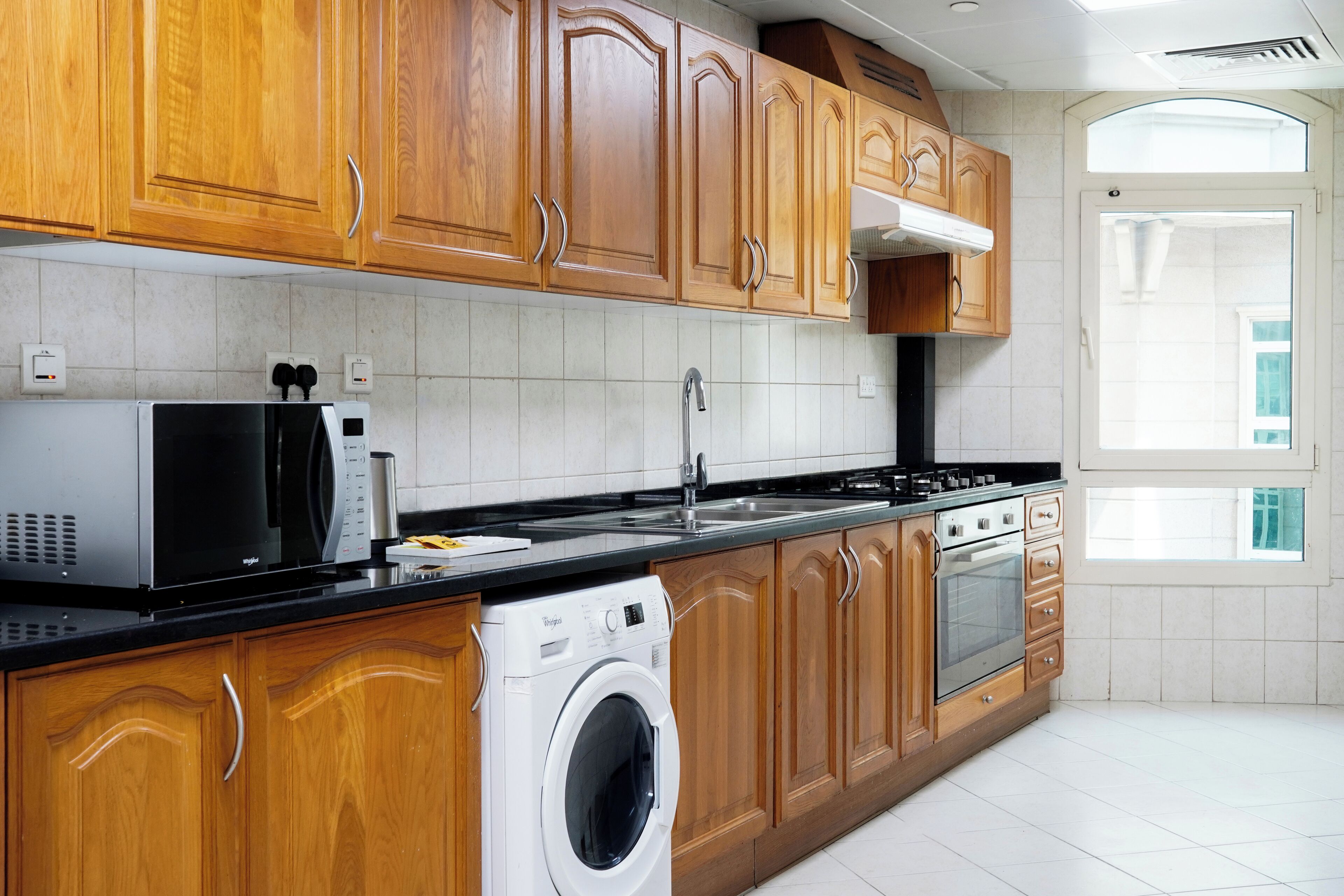 A modern kitchen with wooden cabinets, a stainless steel sink, a microwave, a washing machine, and a built-in oven.