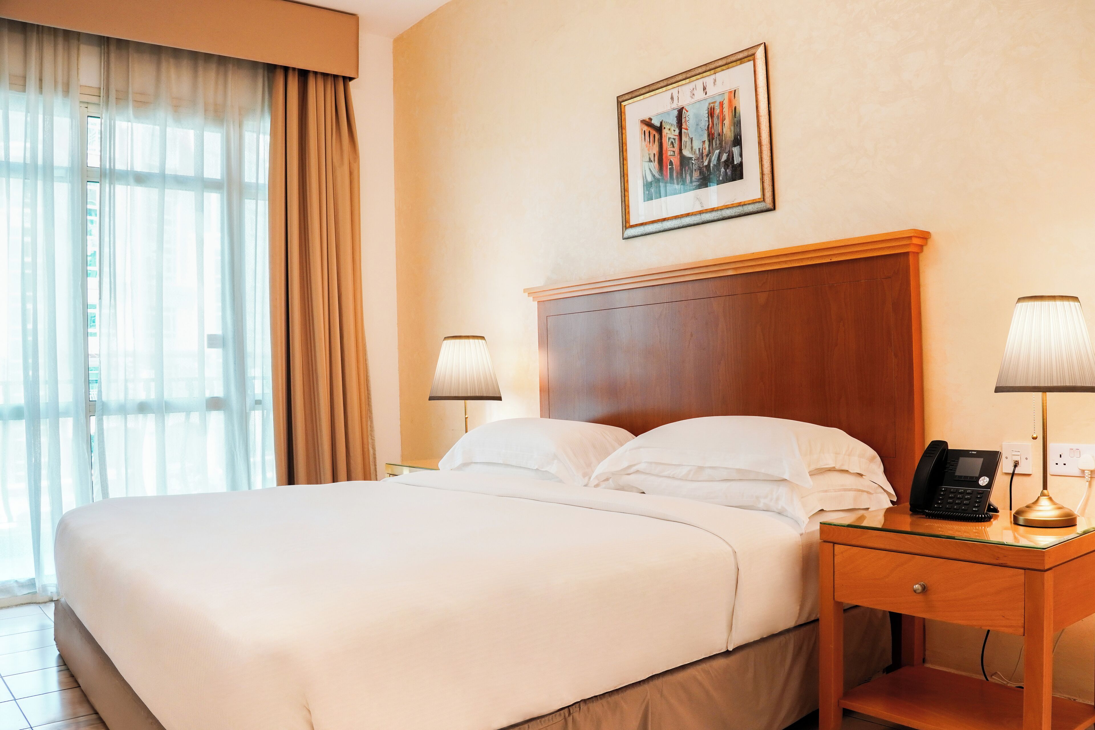 A hotel room with a large bed, two bedside lamps, a wooden nightstand, a framed picture on the wall, and a window with curtains.
