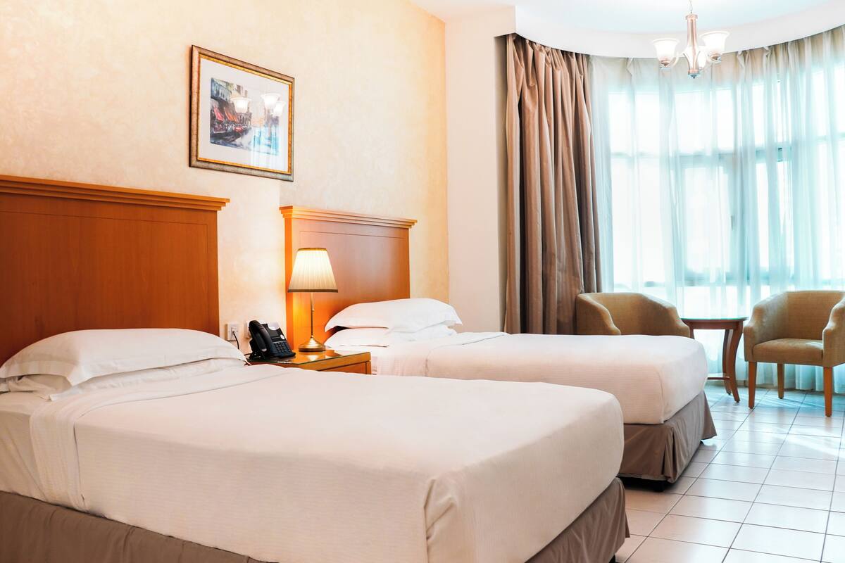 A hotel room with two beds, a bedside table with a lamp, a telephone, a framed picture on the wall, and a window with curtains.