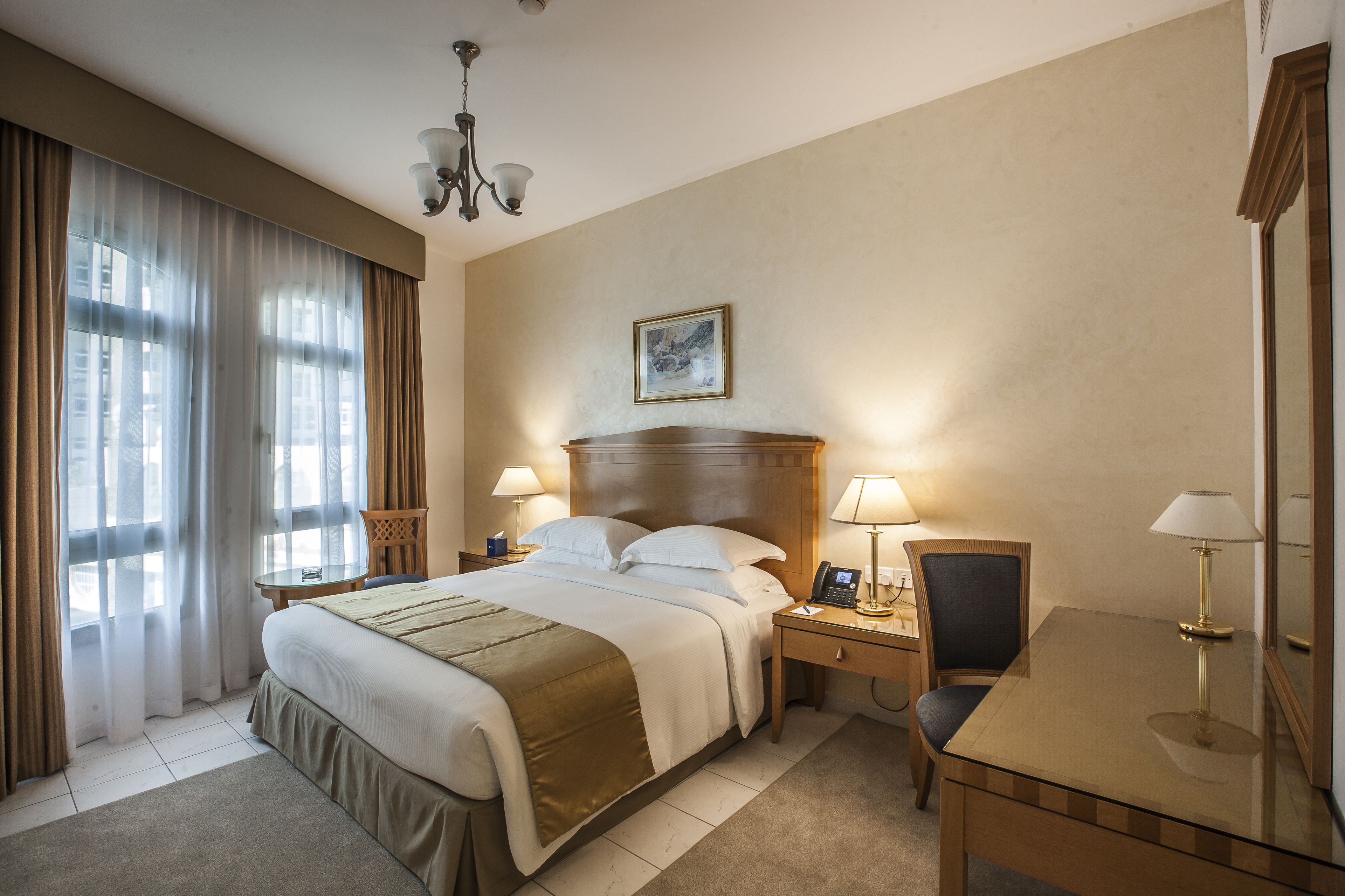 Habitación de hotel con una cama grande, dos mesitas de noche con lámparas, un escritorio con una silla, un candelabro y un cuadro en la pared.