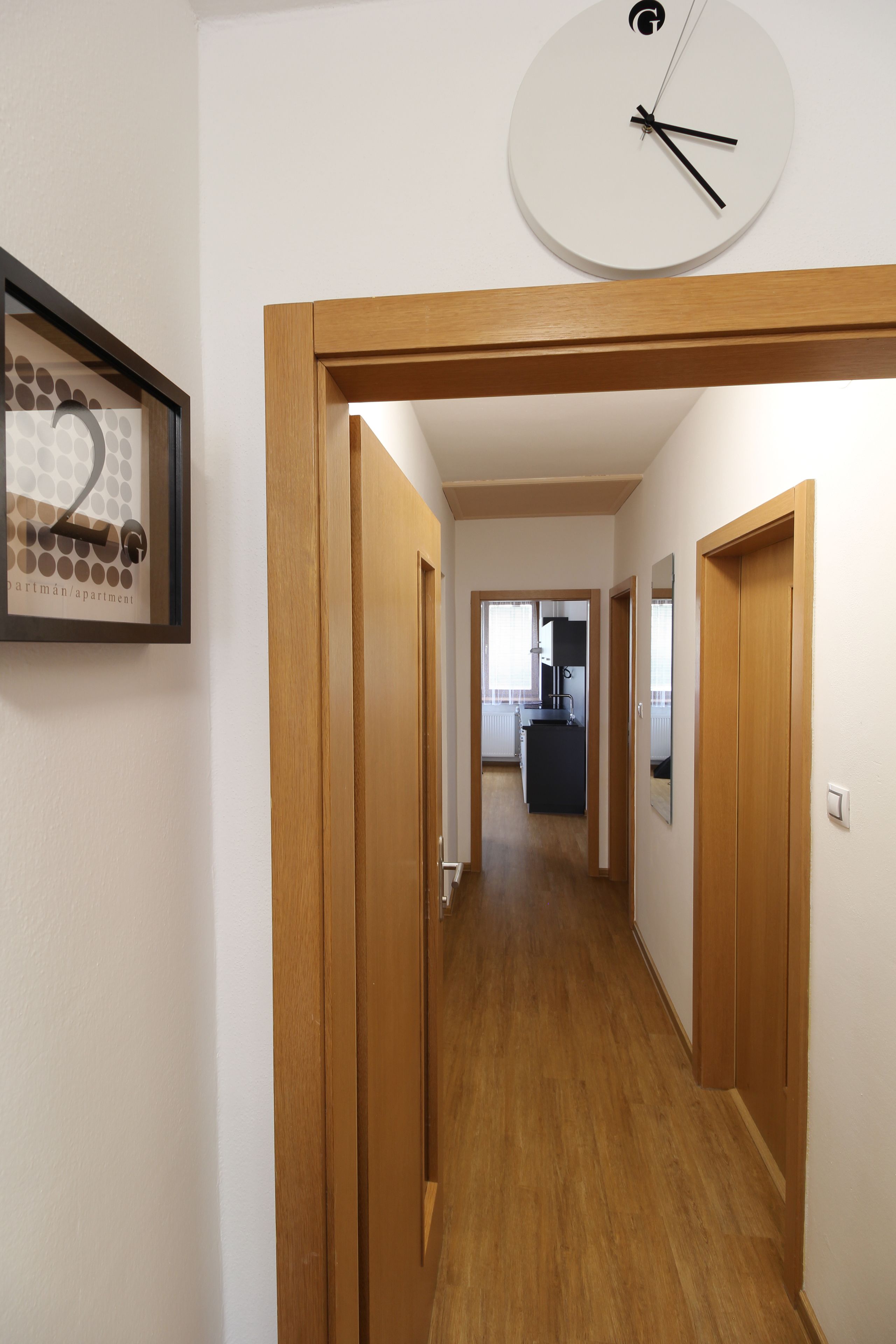 Un couloir avec des portes en bois, un tableau encadré au mur et une horloge.