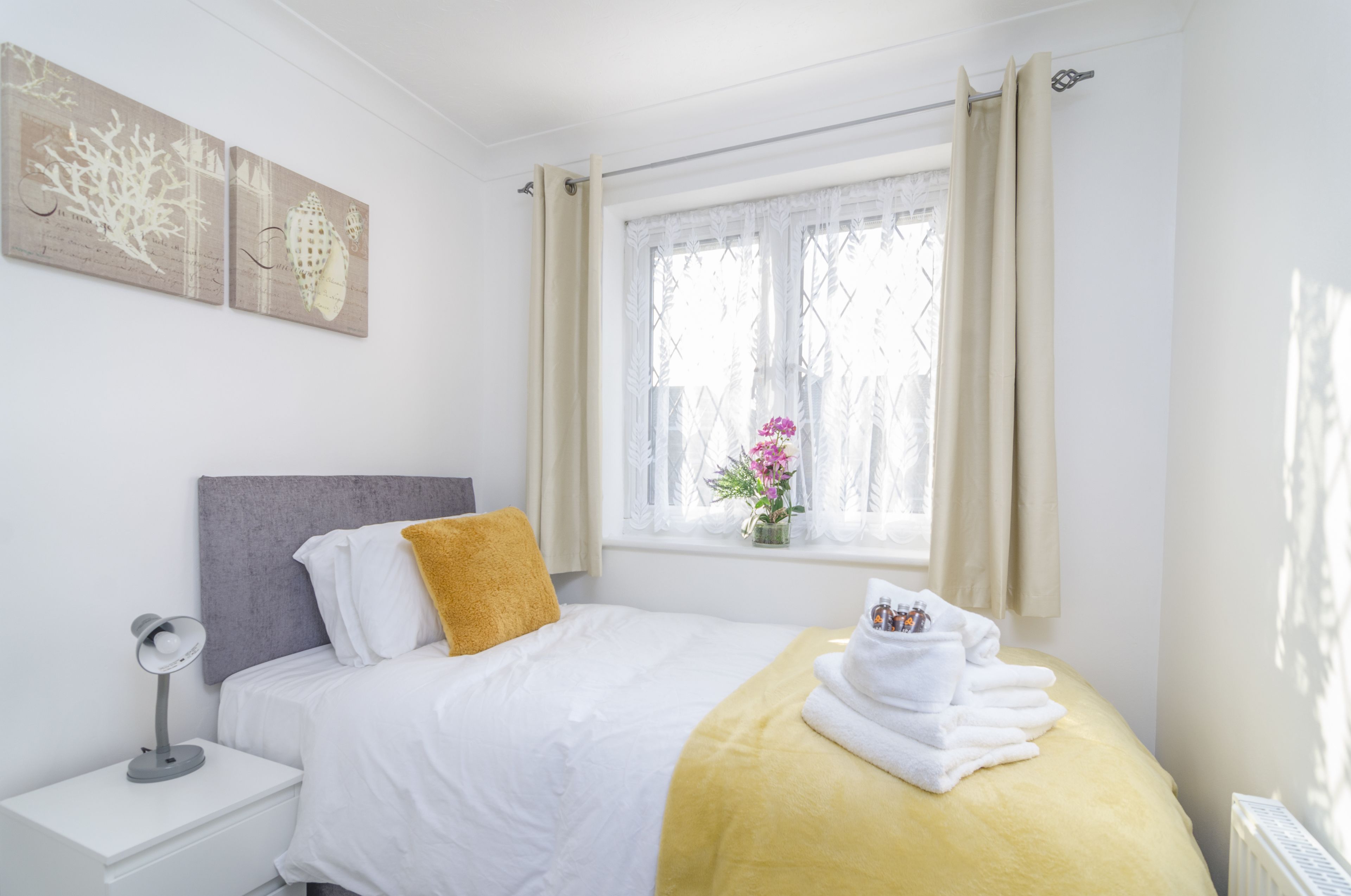 A neatly arranged bedroom with a bed, a bedside table with a lamp, a window with curtains, and a wall with artwork.