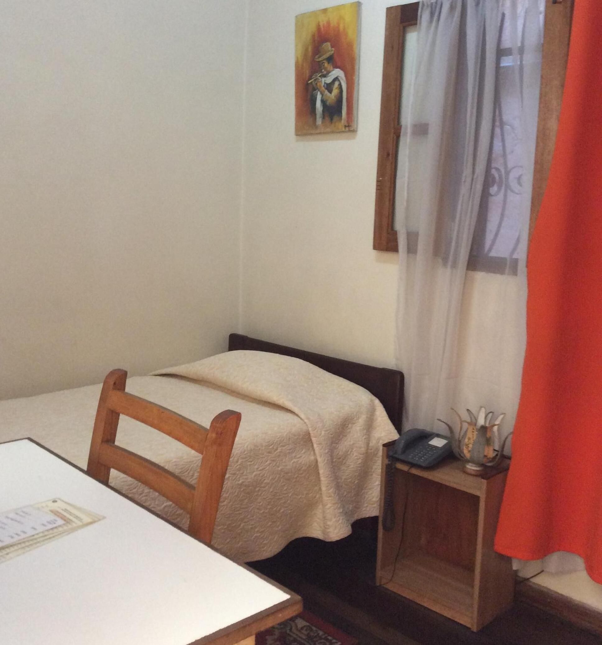 Una stanza piccola e semplice con un letto singolo, una sedia in legno, un tavolino con un libretto dei conti, un telefono e un quadro appeso al muro.