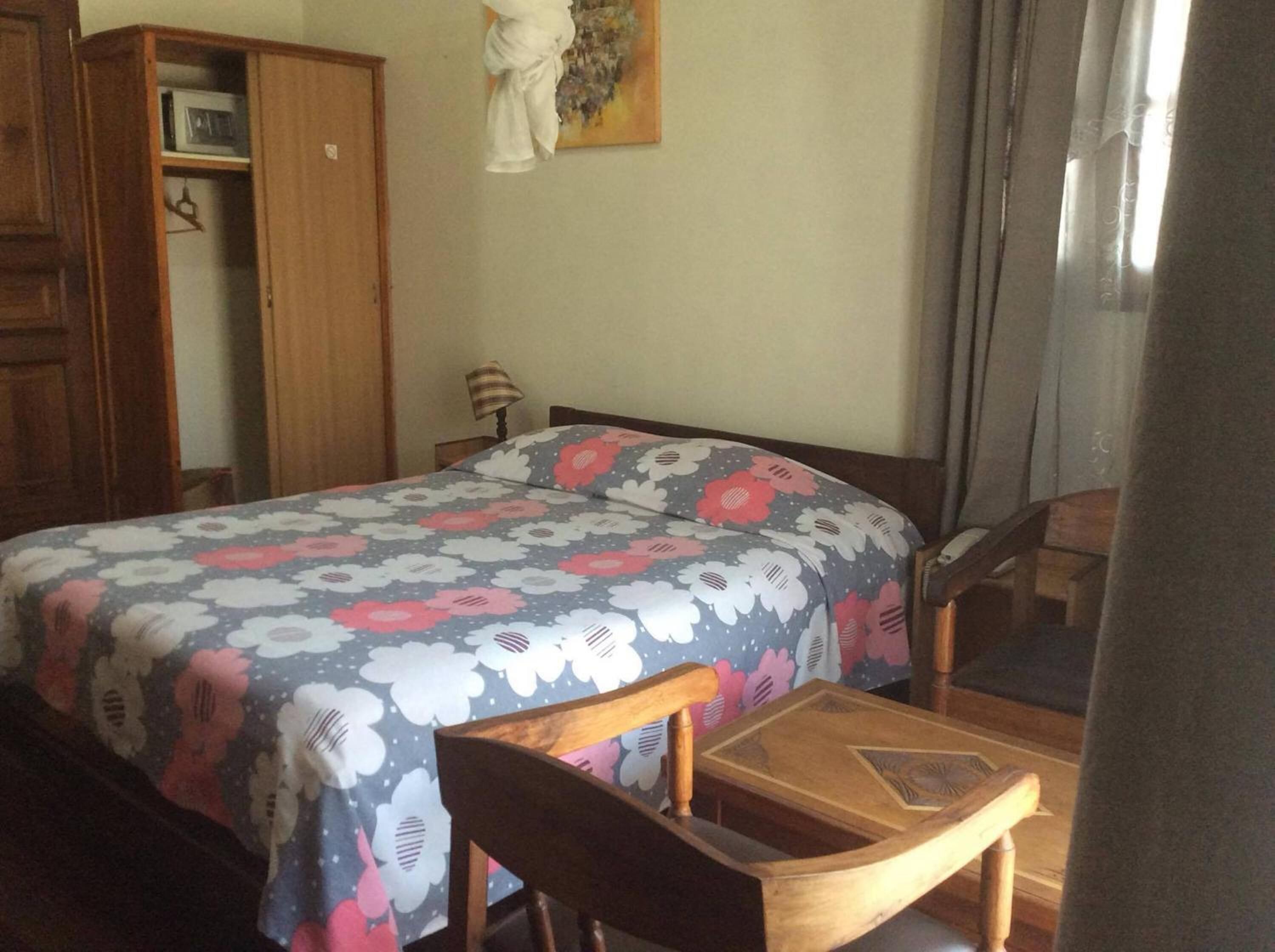 Quarto com cama, cadeira de madeira, mesa pequena, guarda-roupa e um quadro na parede.