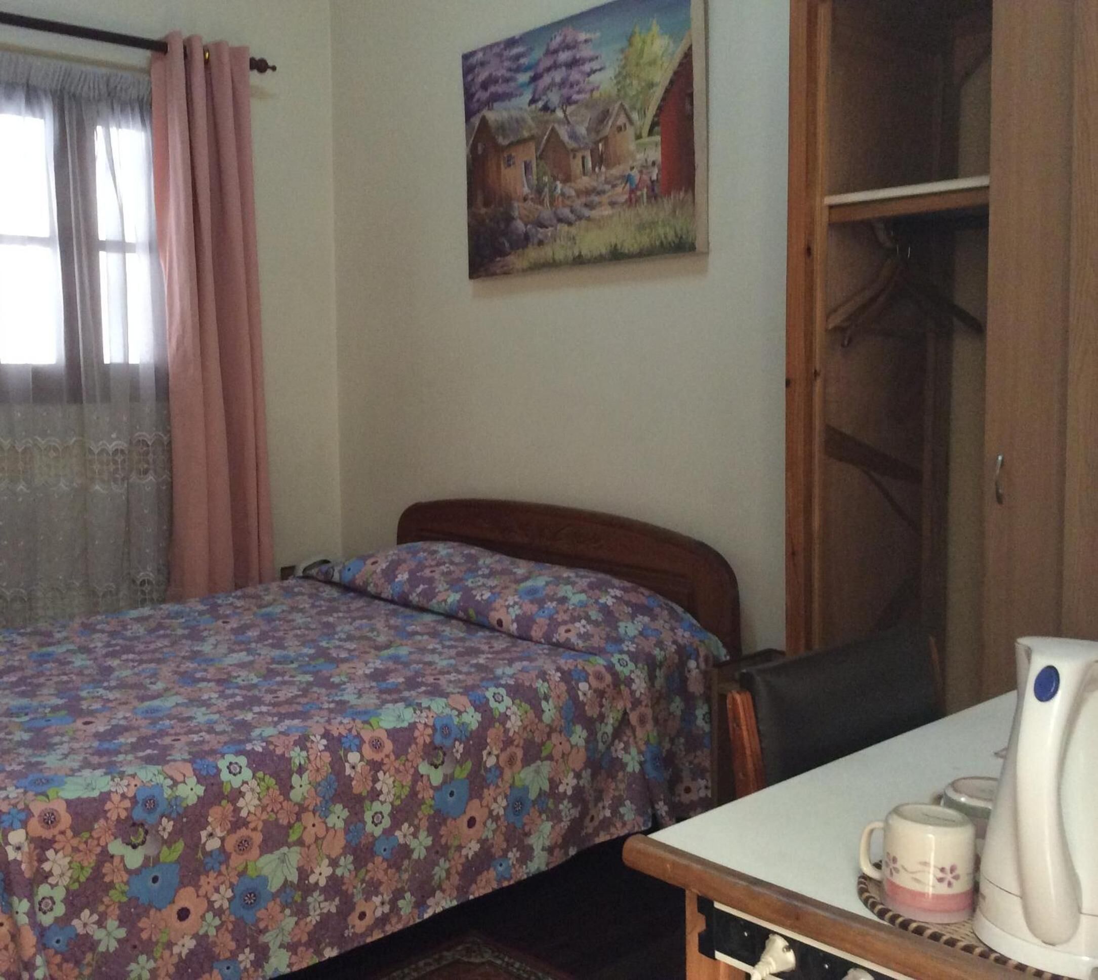 A bedroom with a bed, a chair, a table with a coffee maker, and a painting on the wall.
