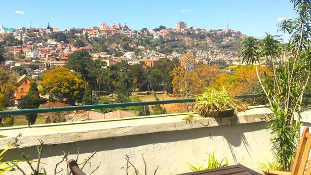 Un balcón con vistas a un paisaje urbano, que incluye una colina con edificios y una torre. Hay plantas en macetas y una mesa de madera con una silla.